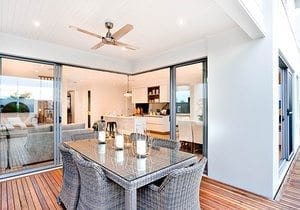 Outdoor patio area with table set up beside an entrance to inside of a modern house with a kitchen, there are candles on the table under the fan